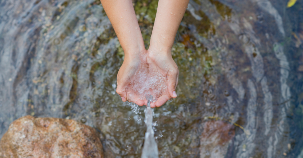 A person’s hands are cupped together, collecting clear flowing water from a natural outdoor source with rocks visible nearby—an image that echoes Malaysia’s hidden water crisis.
