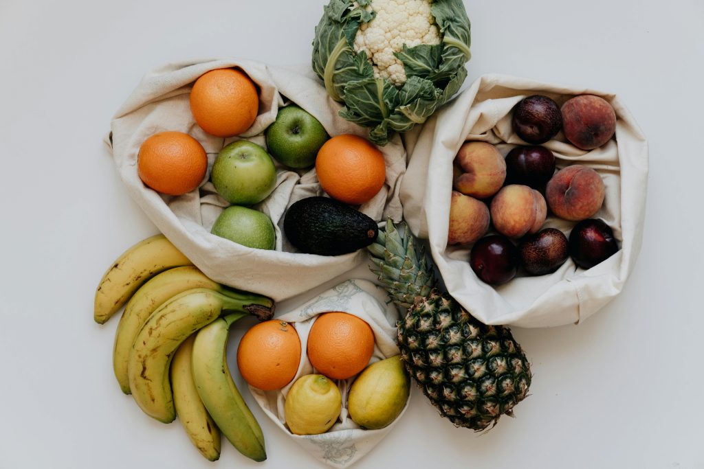 Assorted fresh fruits and vegetables, including oranges, apples, bananas, lemons, avocado, pineapple, peaches, plums, and even a head of ugly vegetables cauliflower—all to help you save money on your grocery bill—arranged on a white surface.