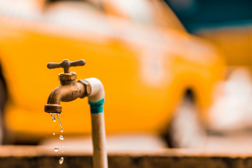 Close-up of a metal outdoor tap with water dripping, attached to a PVC pipe, with a blurred yellow vehicle in the background—reminding scientists of massive hidden water reserves discovered beneath Atlantic depths.