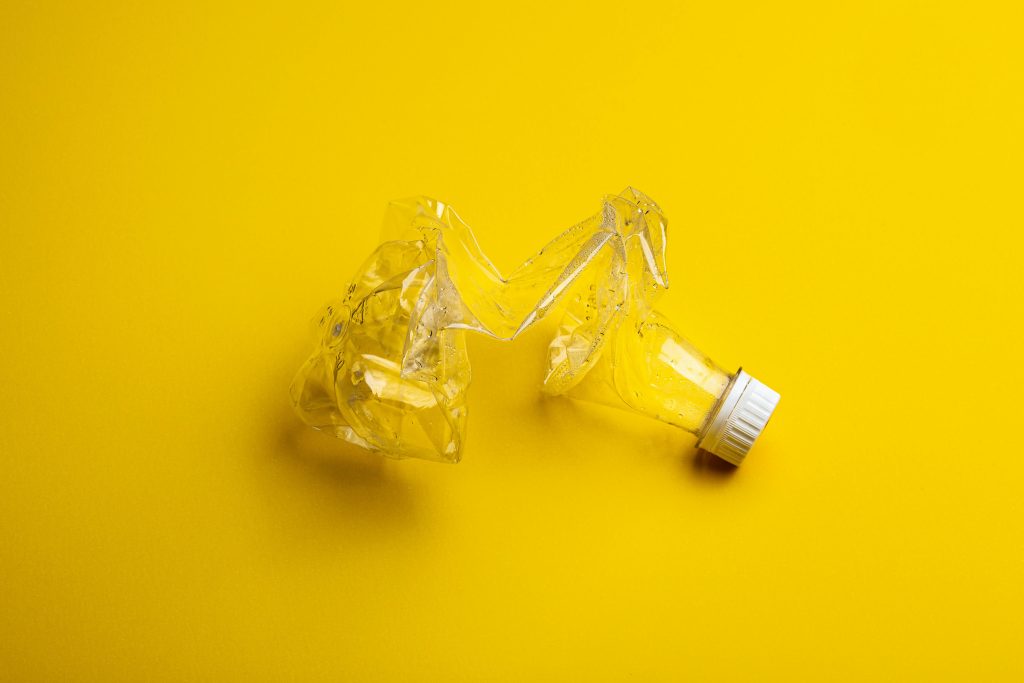 A crushed clear plastic bottle with a white cap lies on a bright yellow background, serving as a subtle reminder of the impact of tourism on the environment.