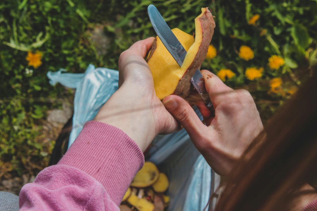 A person peels a potato with a knife outdoors in Scotland, surrounded by green grass and yellow flowers—highlighting simple steps to reduce food waste in everyday life.