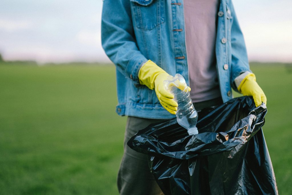 A person wearing yellow gloves and a denim jacket places a plastic bottle into a black garbage bag in a grassy outdoor area, showcasing responsible waste management in the UK.